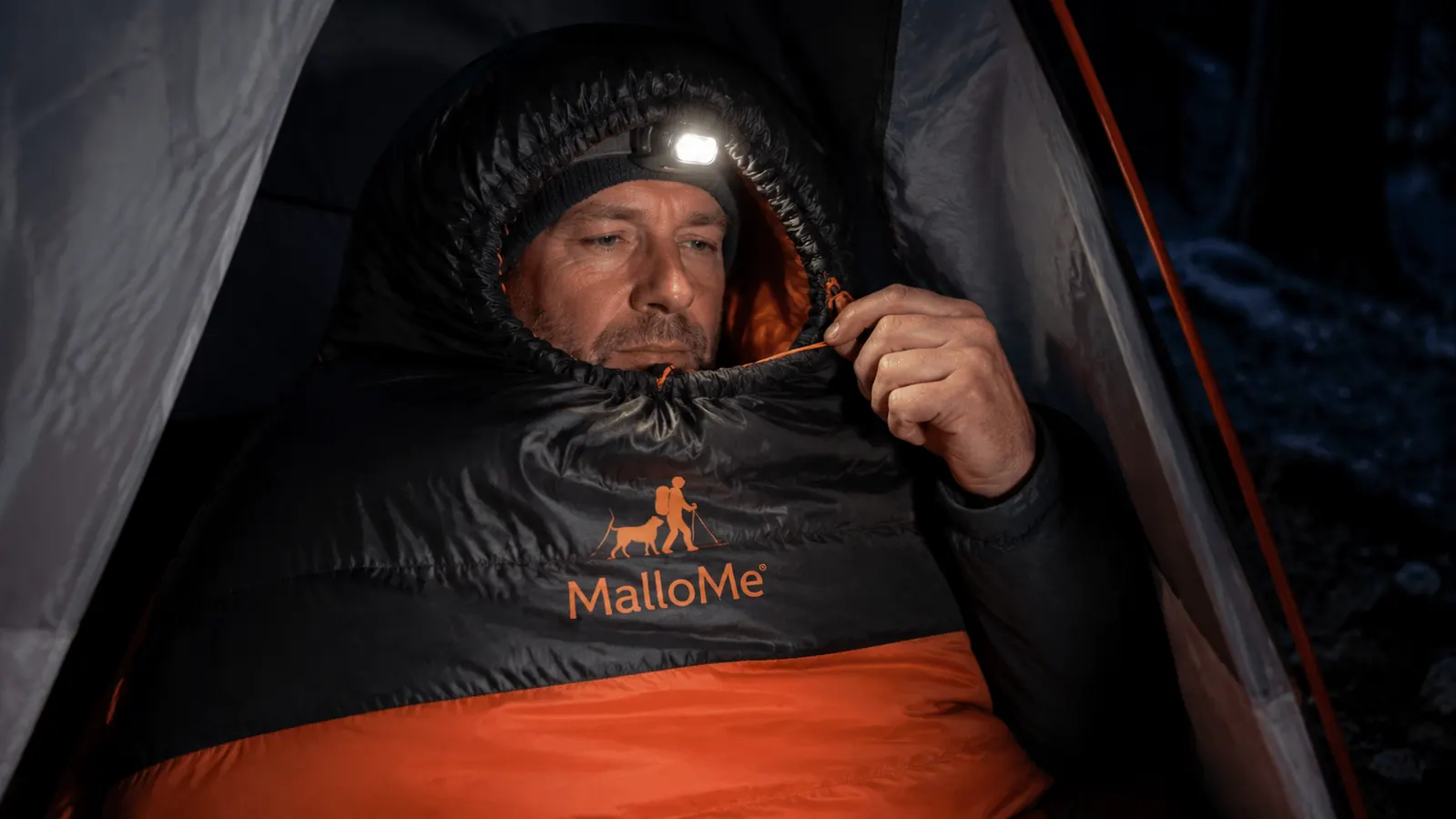 Camper zipping up a mummy sleeping bag inside a dome tent, headlamp on