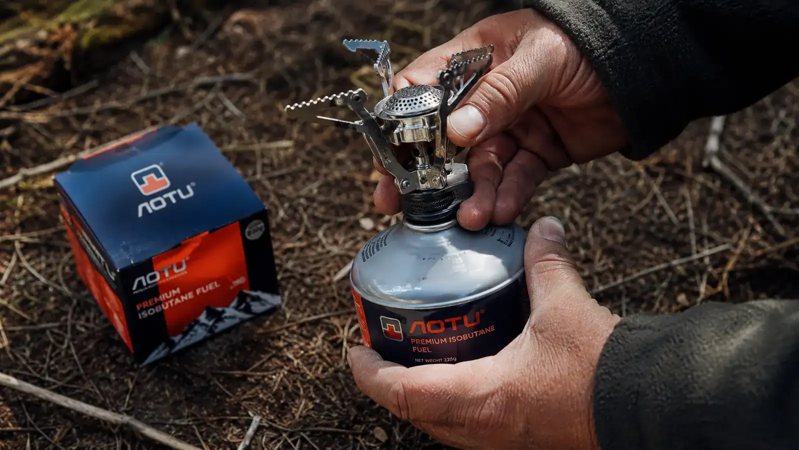 Hand attaching AOTU backpacking stove to an isobutane canister at a trailside campsite