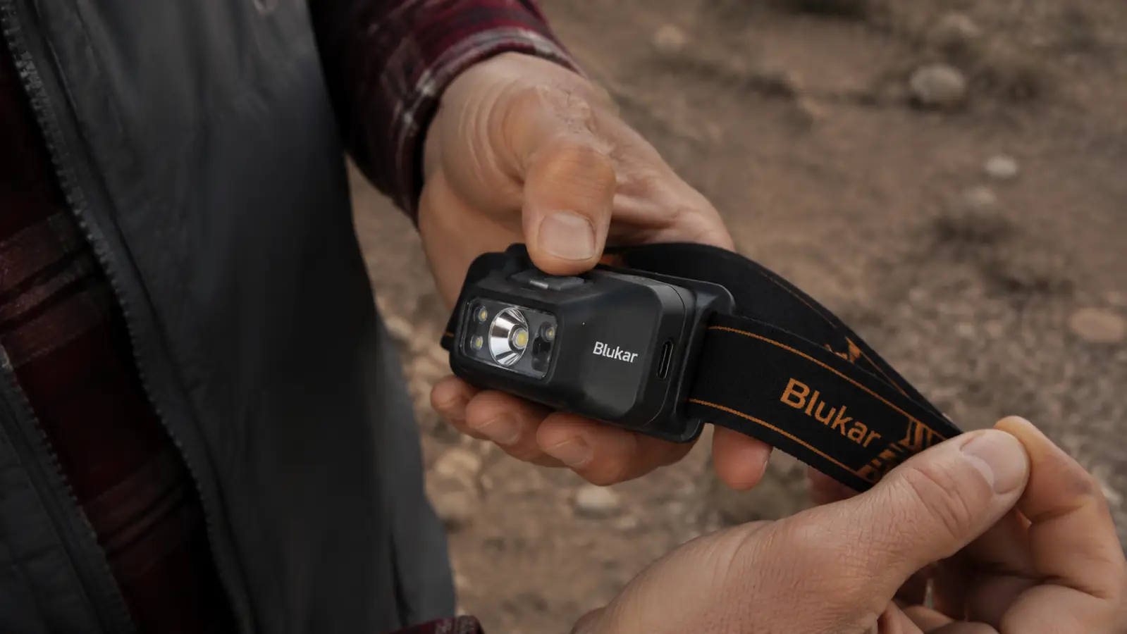 Hand holding a Blukar headlamp showing the USB-C charging port and control button