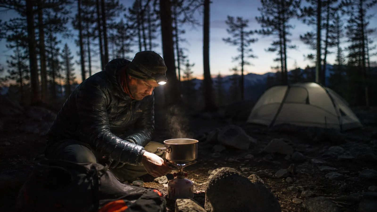 Camper wearing a headlamp while cooking at a camp stove in early morning darkness