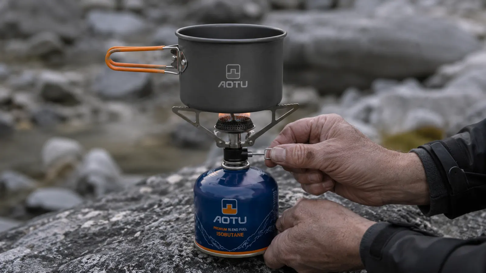 Hands lighting the AOTU backpacking stove on a rocky ledge with a screw-top isobutane canister attached