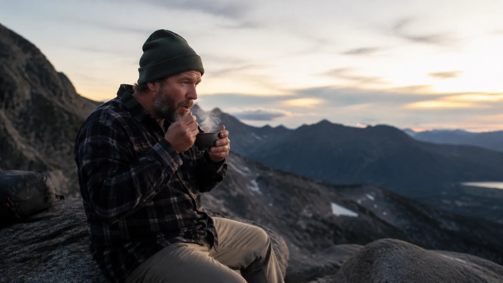Hiker eating a hot meal from a camp cup while sitting on a boulder with mountain ridgelines in the background