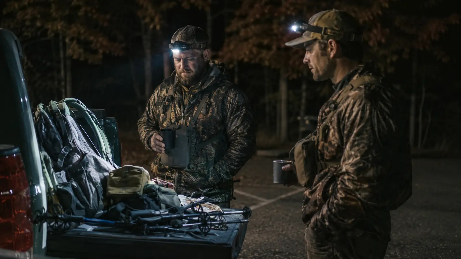 Two hunters at a truck tailgate under headlamp light in a dark forest parking area