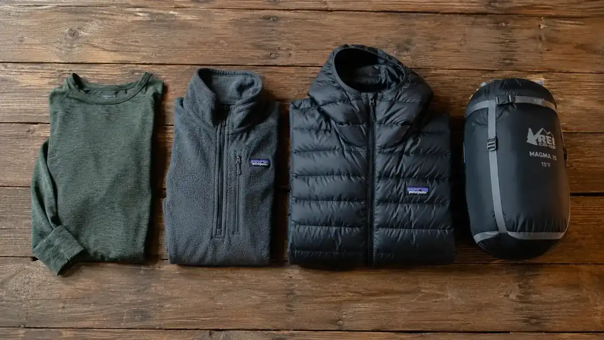 Layered clothing system laid flat showing base layer, mid layer fleece, and insulated jacket next to a sleeping bag
