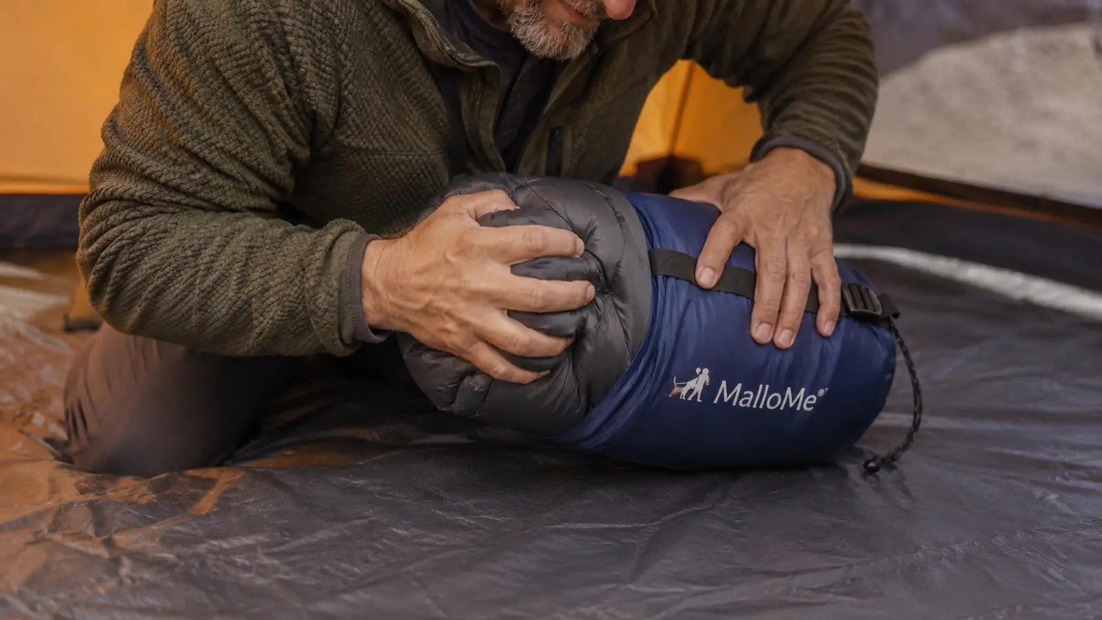 Hands rolling up a compact mummy sleeping bag next to a packed stuff sack on a tent floor
