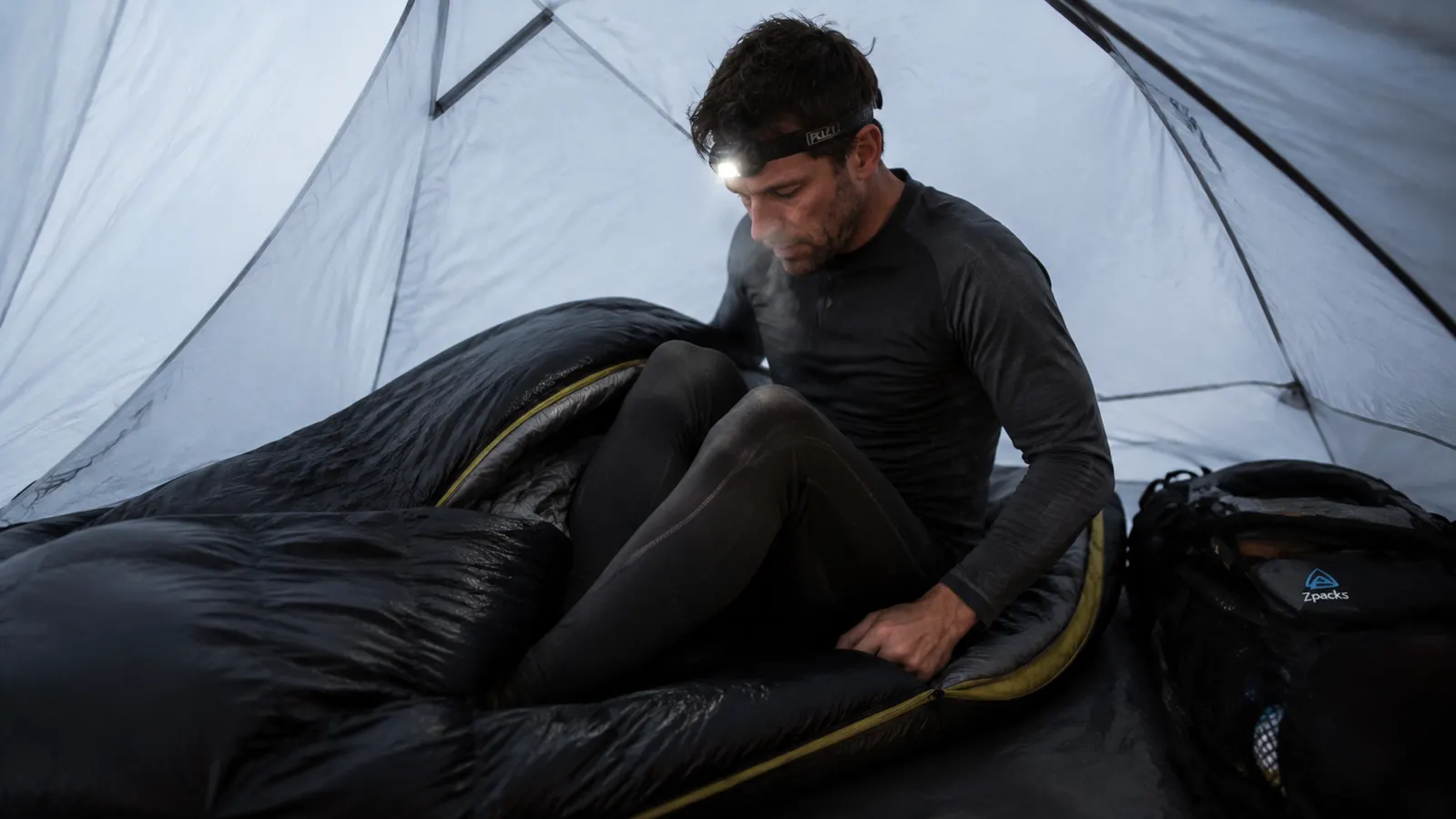 Man in base layers climbing into a mummy sleeping bag in a dark tent, visible breath in cold air